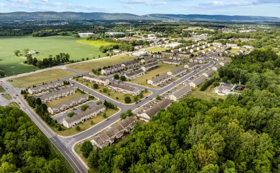 An aerial drone view of the sprawling Keystone Arms community in Carlisle, PA. The image shows several rows of townhomes, residential streets, and vast green spaces. Rolling green hills and distant mountains are visible in the background.