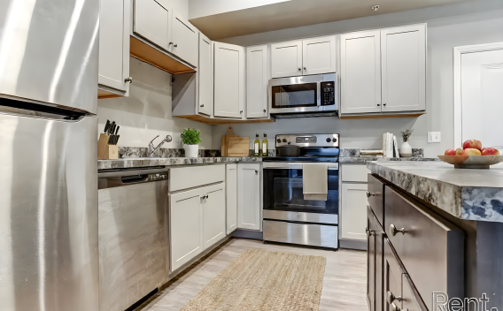 A bright kitchen with stainless steel appliances, including a refrigerator, dishwasher, and stove. The light grey cabinetry is accented by dark grey granite-style countertops and a neutral-toned backsplash. A tan woven rug is centered on the light-colored flooring between the cabinets and a dark wood kitchen island.