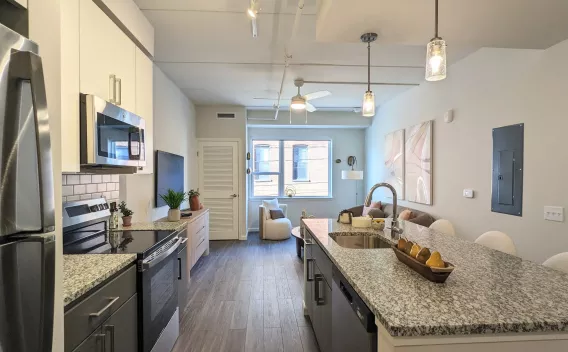 An open-concept layout showing a kitchen with a large granite-style island, stainless steel appliances, and gray lower cabinetry transitioning into the living area.