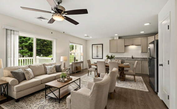 Bright open-plan living room with beige sofa, dining table, and kitchen featuring grey cabinets and stainless steel appliances.