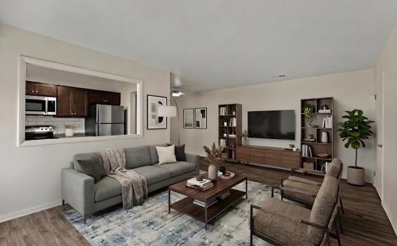 Modern living room with a gray sofa, coffee table, TV, bookshelves, and two armchairs. Kitchen seen through a wall cutout.