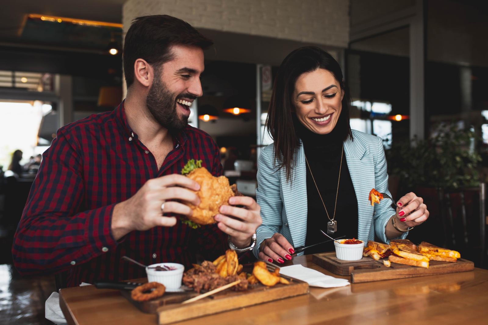 Smiling man and woman enjoying a casual meal together at a restaurant, featuring burgers, fries, and drinks, creating a fun and relaxed dining experience.