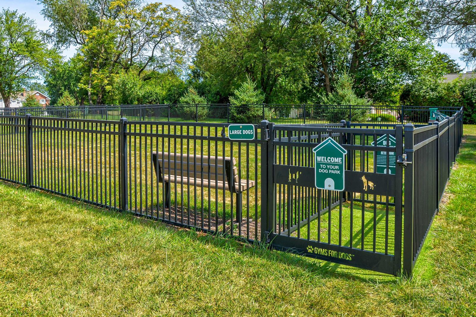 Fenced-in dog park at Village of Olde Hickory Apartments featuring a gated entry, grass play area, and separate zones for large and small dogs, surrounded by mature trees and a park bench.