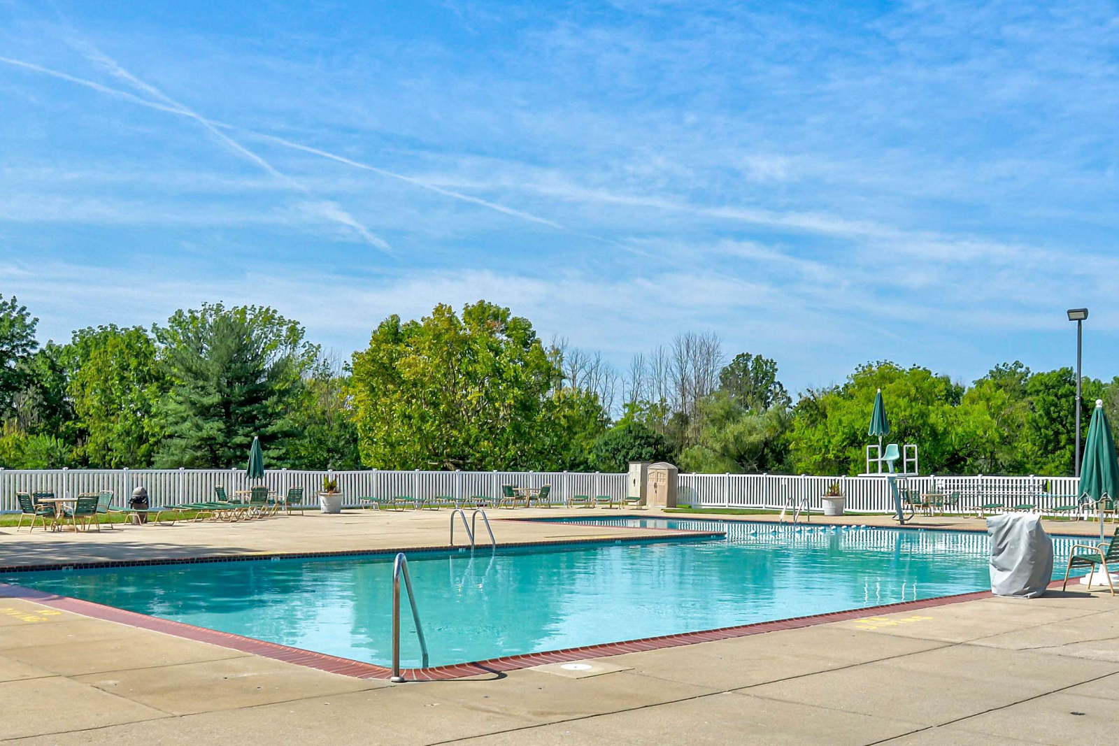 Outdoor rectangular swimming pool at Village of Olde Hickory Apartments, surrounded by lounge chairs, tables with umbrellas, and a white privacy fence with trees in the background under a bright blue sky.