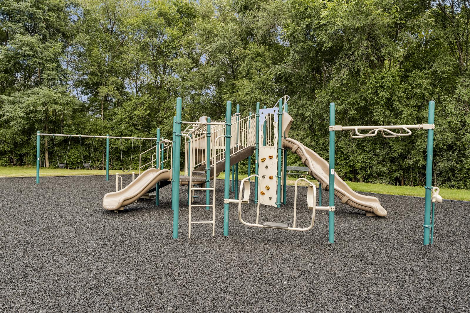 A view of the community playground at Keystone Arms in Carlisle, PA, featuring swings, slides, climbing equipment, and a large, mulched play area surrounded by mature trees. 