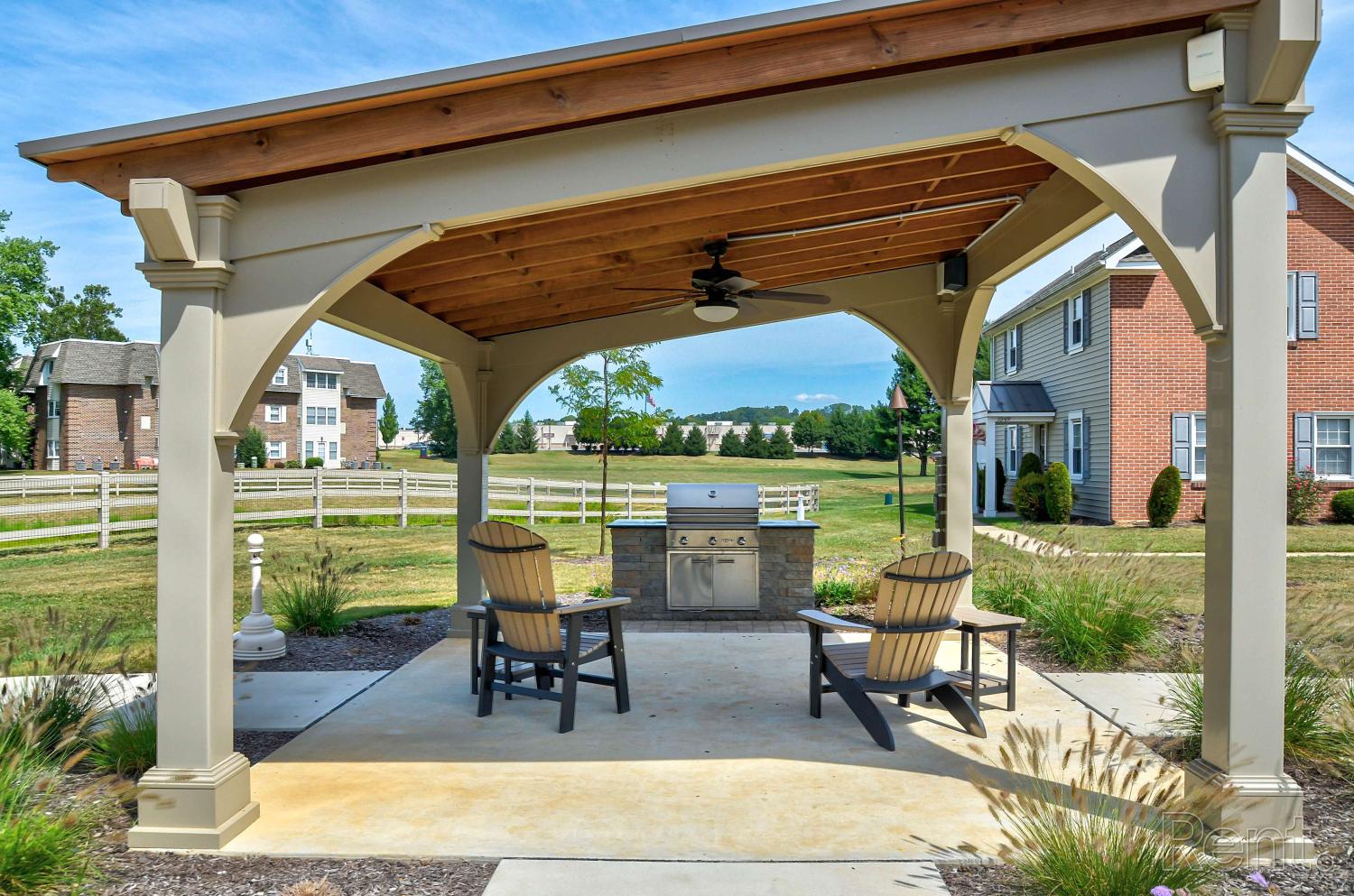 Covered outdoor pavilion with Adirondack chairs, ceiling fan, and built-in stainless steel grill surrounded by landscaped greenery and open lawn.