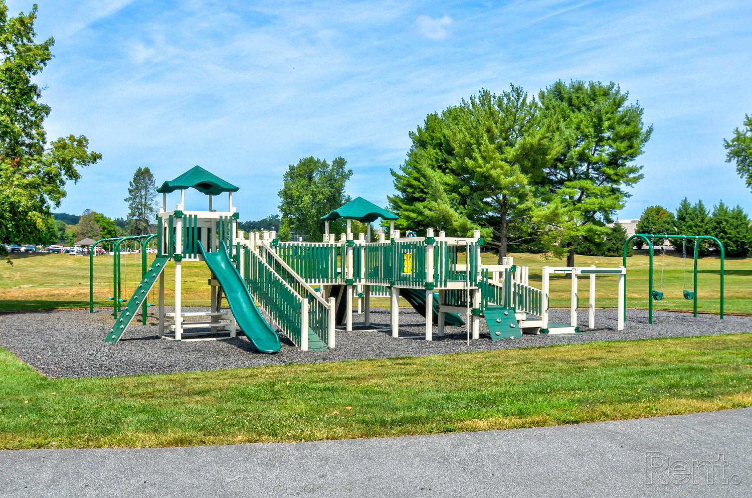 Large outdoor playground with green slides, climbing structures, and swings set on a mulch surface, surrounded by grassy fields and mature trees.