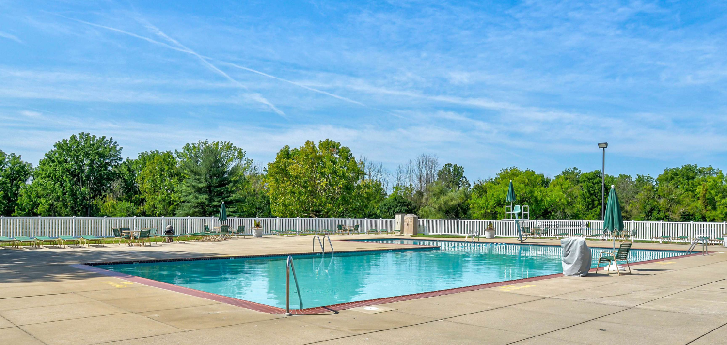 Outdoor rectangular swimming pool at Village of Olde Hickory Apartments, surrounded by lounge chairs, tables with umbrellas, and a white privacy fence with trees in the background under a bright blue sky.