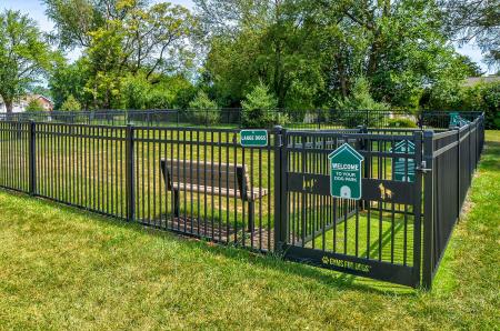 Fenced-in dog park at Village of Olde Hickory Apartments featuring a gated entry, grass play area, and separate zones for large and small dogs, surrounded by mature trees and a park bench.