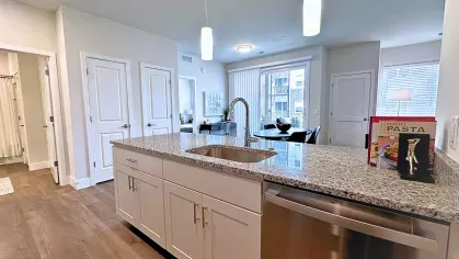 Bright open-concept layout with granite kitchen island, double sink, stainless steel dishwasher, and a view into the living and dining spaces.