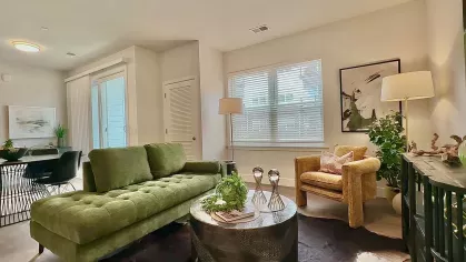 Furnished living space with green and mustard seating, large windows with blinds, and layered decor including plants, wall art, and a circular coffee table.