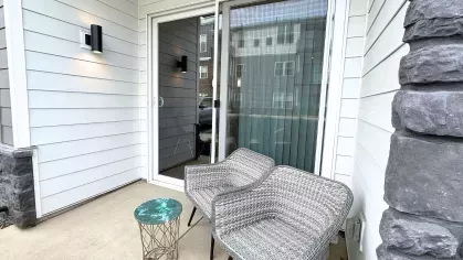 Cozy private patio with two woven chairs and a side table, bordered by siding and stone detailing, accessed through sliding glass doors.
