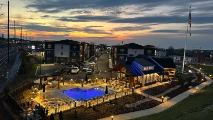 Nighttime aerial view of The Foundry community with the illuminated pool, clubhouse, and American flag under a glowing sunset sky.