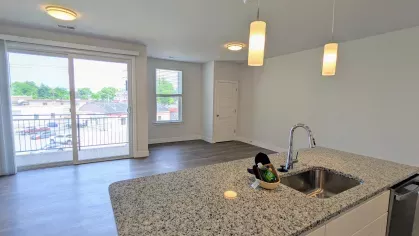 Open-concept kitchen featuring a granite island with a sink, pendant lighting, and a view into the living area with large windows and balcony access.