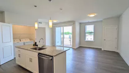 Spacious open-concept layout featuring a modern kitchen island with granite countertops, pendant lighting, and in-unit laundry area next to large windows and balcony access.
