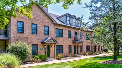 Red brick apartment building with classic architecture, green doors, and a shaded pathway lined with trees and shrubs.