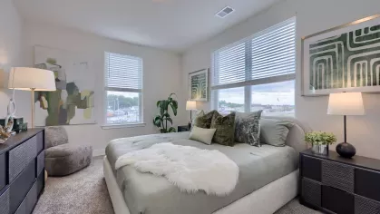 Bright and spacious bedroom featuring large windows with blinds, a cozy bed with green-toned bedding, modern abstract artwork, and soft carpet at The Forge apartments.