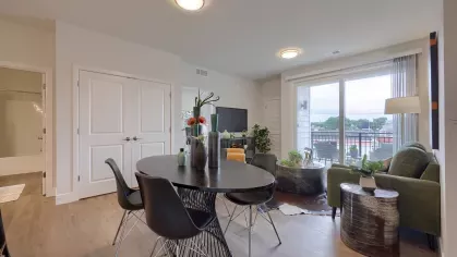 Open-concept living and dining area with round dining table, modern black chairs, and access to balcony through sliding glass doors at The Forge apartments.