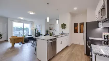 Stylish kitchen with granite countertops and stainless appliances, opening into a cozy living area with a mustard accent chair at The Forge apartments.