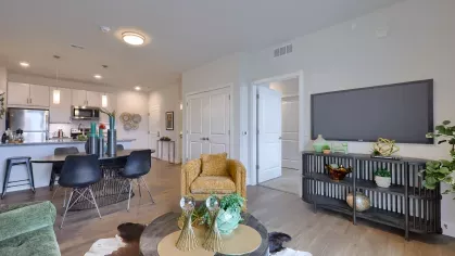 Stylish open-concept living space with green and gold accent furniture, flat-screen TV, and view into the modern kitchen and dining area.