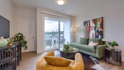 Modern living room with plush seating, indoor plants, and large sliding glass doors opening to a private balcony with outdoor seating.