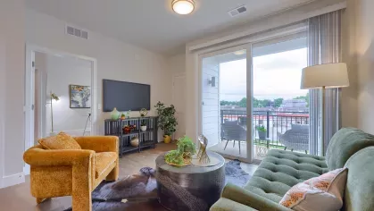 Bright apartment interior with dining set, art-filled living room, and sliding glass doors leading to a furnished balcony.