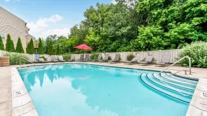 Spacious resort-style pool with entry steps, red umbrellas, and multiple lounge chairs, set against a backdrop of greenery and townhomes.