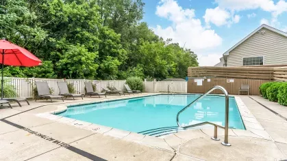 Bright view of community pool area with adjacent clubhouse, poolside seating, red umbrella, and well-maintained landscaping.