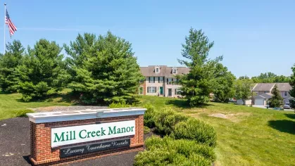 Mill Creek Manor brick entrance sign with bold green lettering and manicured shrubbery, framed by mature pine trees and blue sky.