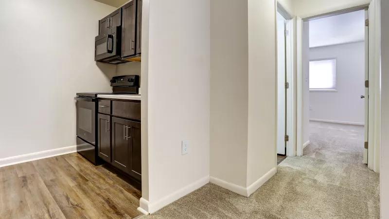Hallway in a renovated Rosedale apartment with carpet leading to a bedroom, adjacent to the kitchen with dark wood cabinets and appliances.