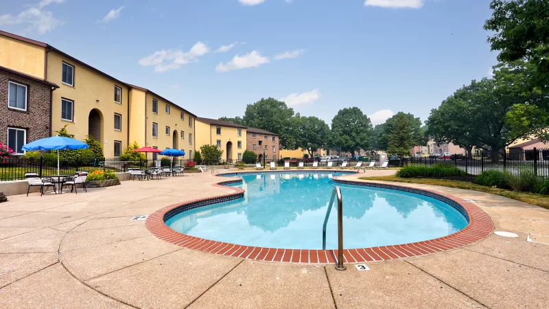 Refreshing, freeform swimming pool at Rosedale, featuring clear blue water, a brick border, lounge chairs, and apartment buildings in the background.