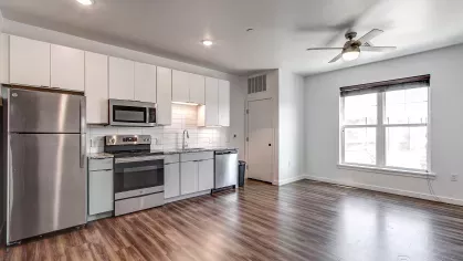 Open layout apartment at Stadium Row highlighting the modern kitchen with white cabinets and stainless steel appliances, seamlessly connecting to the living area with a ceiling fan and large window.