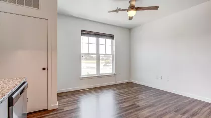 Living space in an open-plan apartment at Stadium Row, featuring wood-style flooring, a ceiling fan, and a large window providing natural light.
