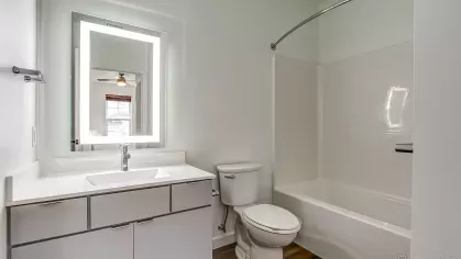 Modern bathroom in the Slugger 208 floor plan at Stadium Row, featuring a white vanity with an illuminated mirror, toilet, and a bathtub with shower.