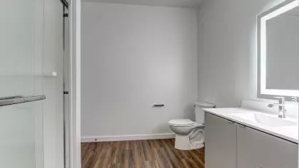 Sleek bathroom in the Slugger 208 floor plan at Stadium Row, featuring a glass-enclosed shower, modern vanity with illuminated mirror, and wood-style flooring.