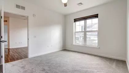 Bedroom in the Slugger 208 floor plan at Stadium Row with gray carpet, a ceiling fan, and a large window overlooking the vibrant community.