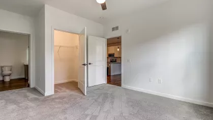 Bedroom in the Slugger 208 floor plan at Stadium Row, featuring soft gray carpet, a ceiling fan, and open doorways leading to a closet and the modern kitchen.
