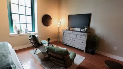 Living space with large industrial-style window, two green chairs, flat-screen TV, and modern dresser at Prince Street Lofts