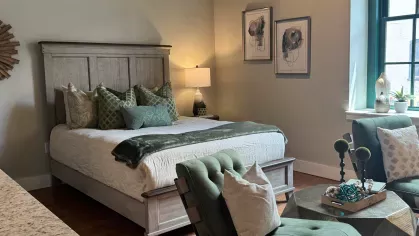 Studio bedroom setup with wood bed frame, green accent pillows, and seating area near natural light window at Prince Street Lofts
