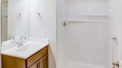A clean and modern bathroom at Riverview Terrace, complete with a spacious vanity with a wood-finish cabinet, white sink, large mirror, and a sparkling white full-size shower.