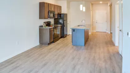 Expansive view of an open concept apartment floor plan at Riverview Terrace, showcasing a contemporary kitchen with dark cabinetry, black appliances, a large island, and ample living space with light wood-style flooring.