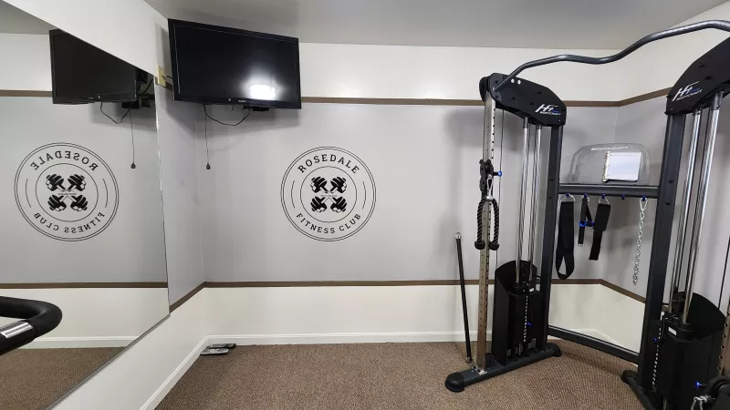 Well-equipped fitness center at Rosedale with strength training machines, free weights, a mounted TV, and a "Rosedale Fitness Club" logo on the wall.