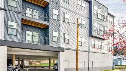 Side exterior view of the Riverview Terrace apartment building, highlighting its modern architecture, private balconies on upper floors, and convenient covered parking accessible to residents on the ground level.