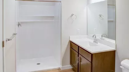 A contemporary bathroom at Riverview Terrace featuring a stylish wood-tone vanity, crisp white countertop with sink, large mirror, and a spacious white stand-up shower with modern fixtures.