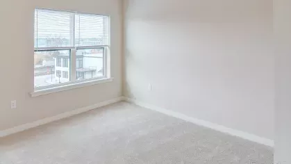 A serene and bright bedroom at Riverview Terrace, featuring soft carpeting, neutral walls, and a large window with blinds that allows for ample natural light, creating a peaceful and inviting living space.