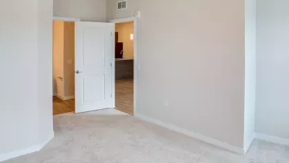 A bright and spacious bedroom at Riverview Terrace, featuring plush carpeting and neutral walls, with open doorways revealing the functional apartment layout leading to other modern living spaces.