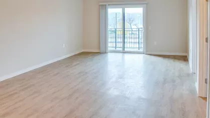 A spacious living room at Riverview Terrace featuring durable wood-style flooring, neutral walls, and a large sliding glass door with blinds leading to an outdoor balcony, providing abundant natural light.