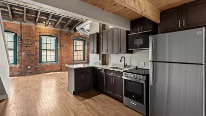 Modern kitchen and living space with exposed brick walls, original wood beam ceiling, stainless steel appliances, and large loft-style windows.