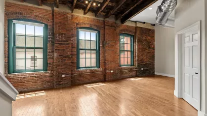 Spacious living room with hardwood flooring, three large arched windows, and original brick interior walls for a classic urban loft aesthetic.