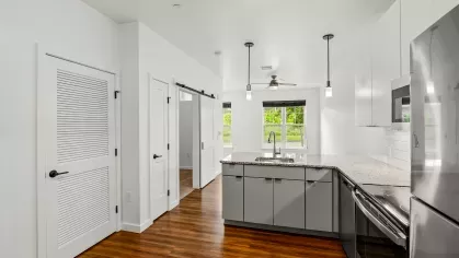 Modern apartment kitchen at Stadium Row featuring stainless steel appliances, granite countertops, sleek white cabinetry, and wood-style flooring with natural light from adjacent living area
